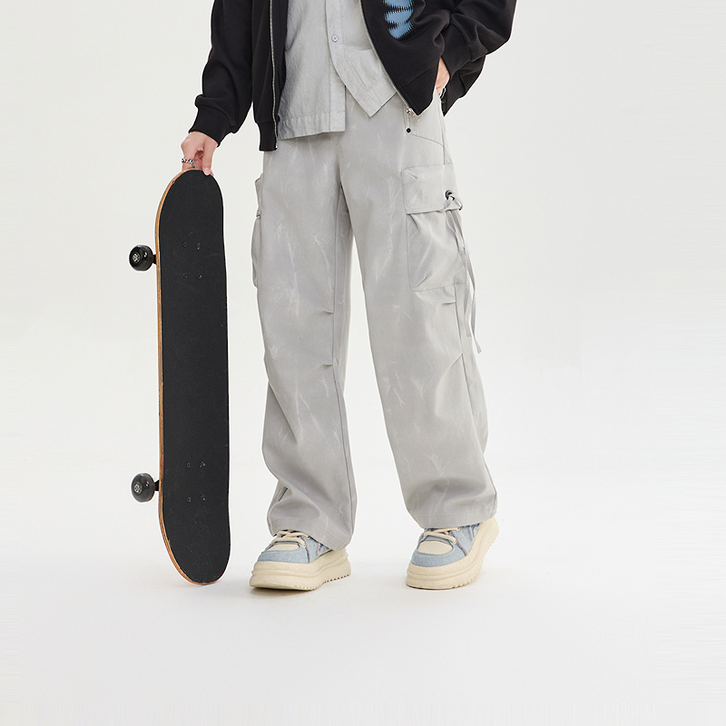 Person holding a skateboard wearing light gray cargo pants and a black jacket on a white background
